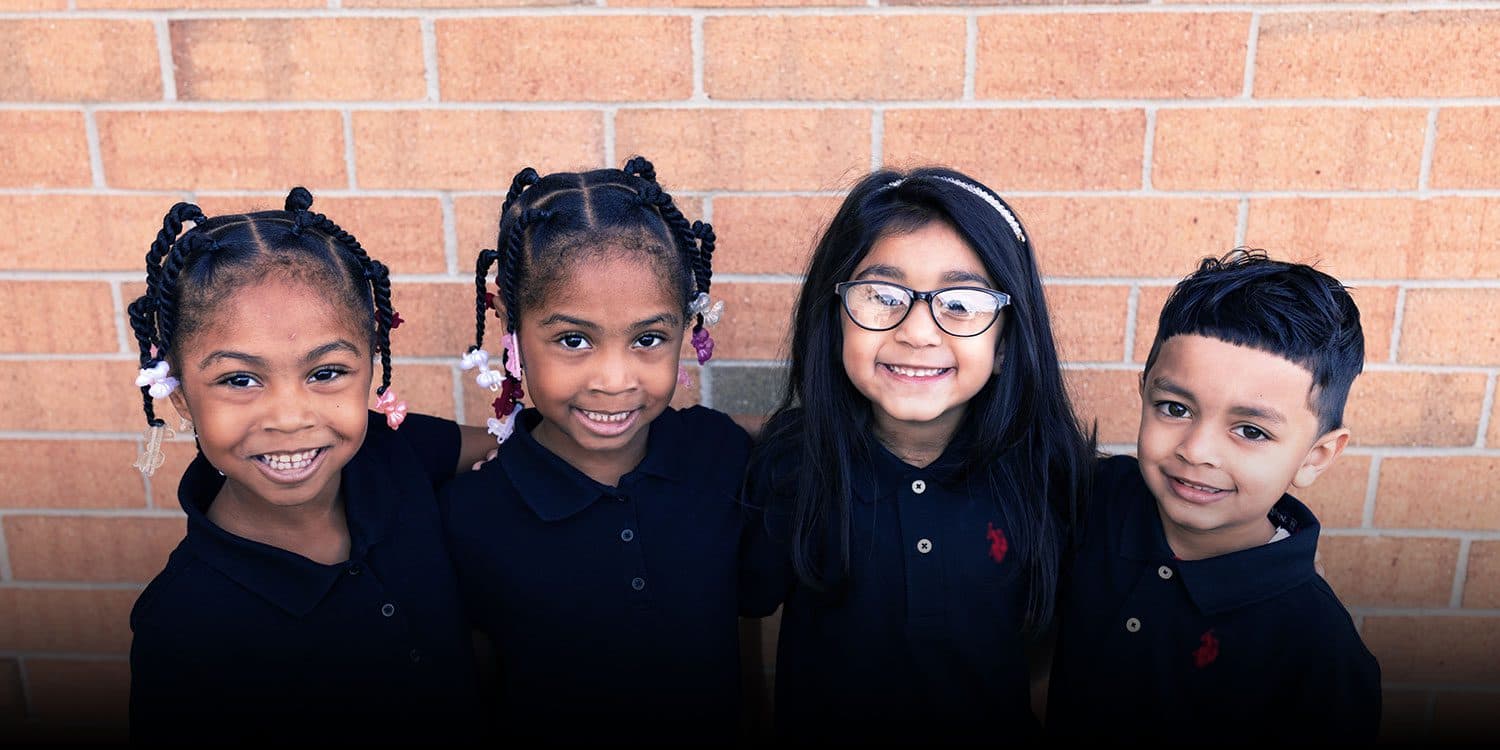 Four smiling students standing together against a brick wall.