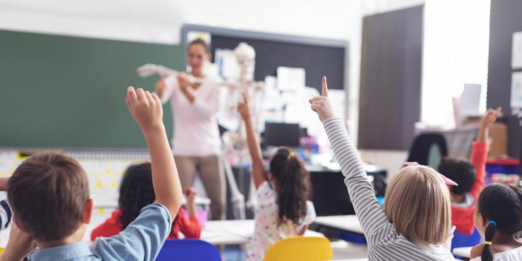 Teacher in front of elementary classroom teaching a science lesson and students with hands raised.